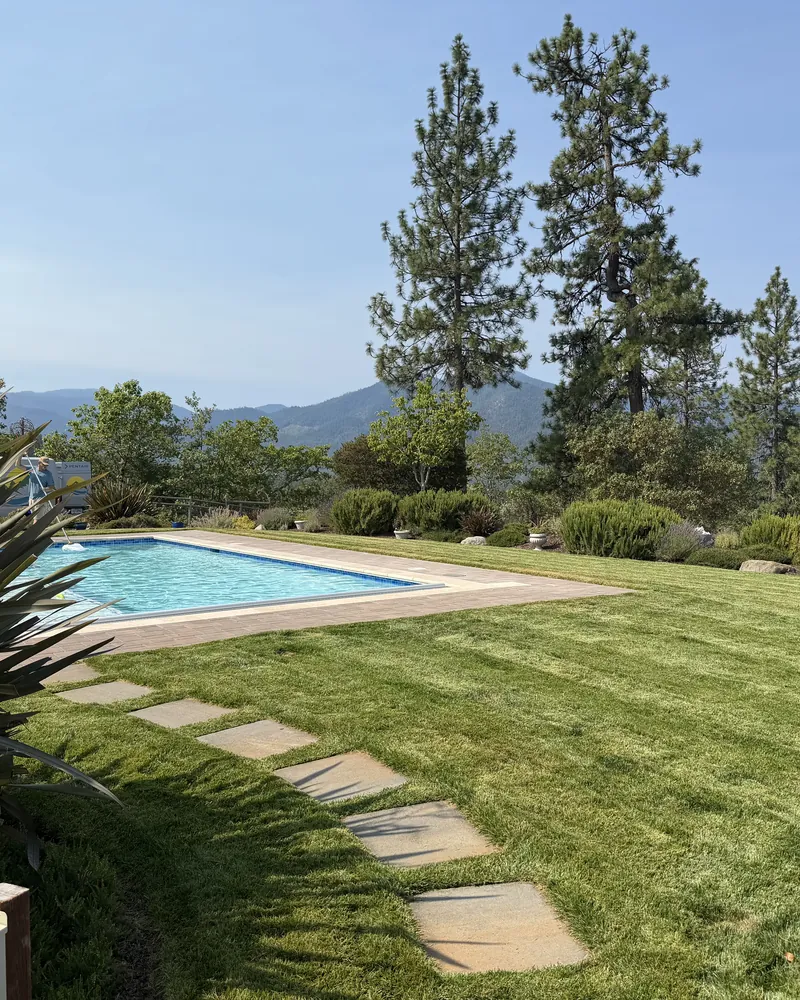 Manicured lawn, flagstone stepping stone path and trimmed shrub beds around a backyard pool with Southern Oregon hills behind